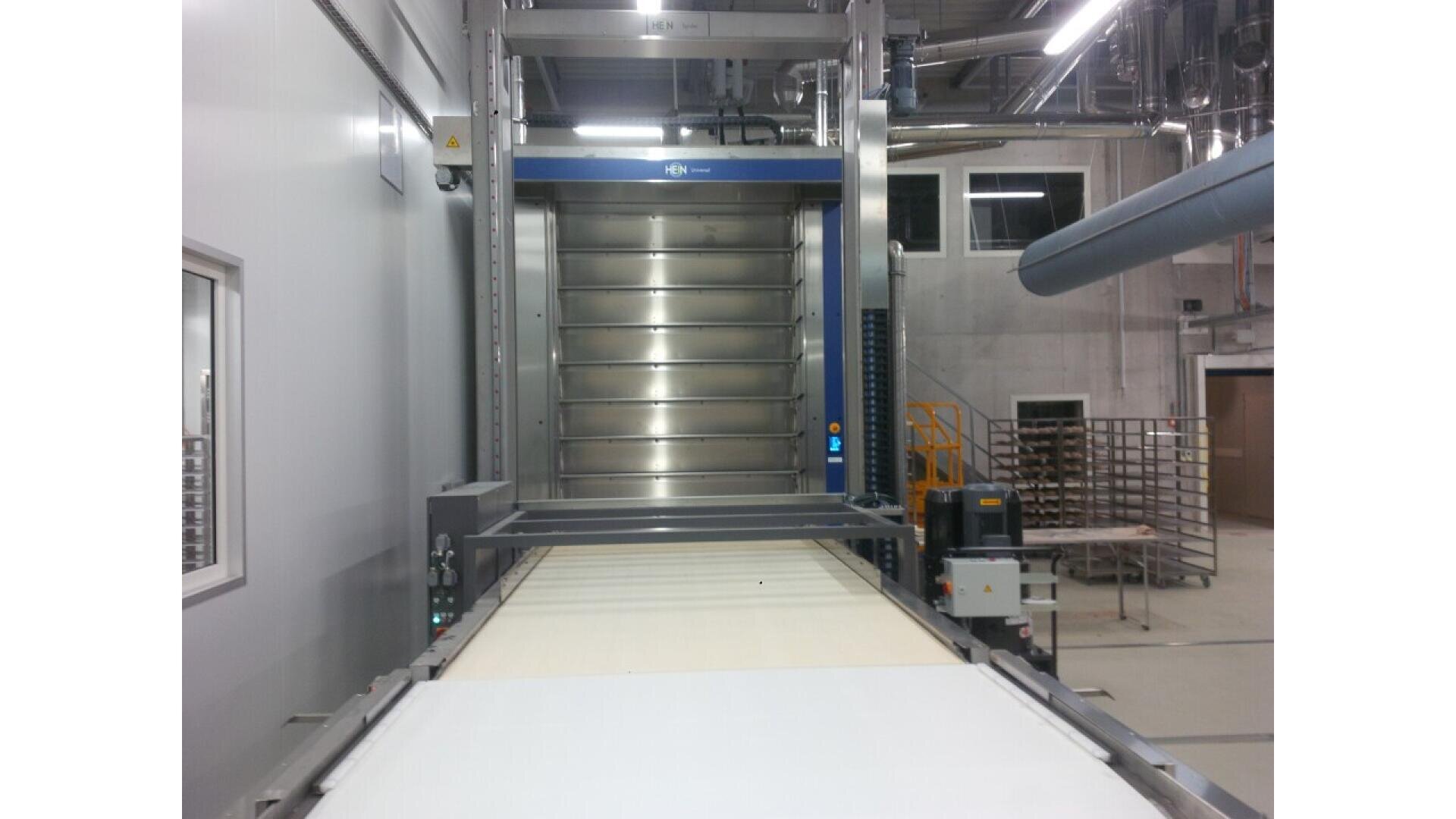A large industrial bread oven with multiple stacked baking chambers stands at the end of a conveyor belt in a modern commercial bakery. Metal racks and equipment are visible in the background.