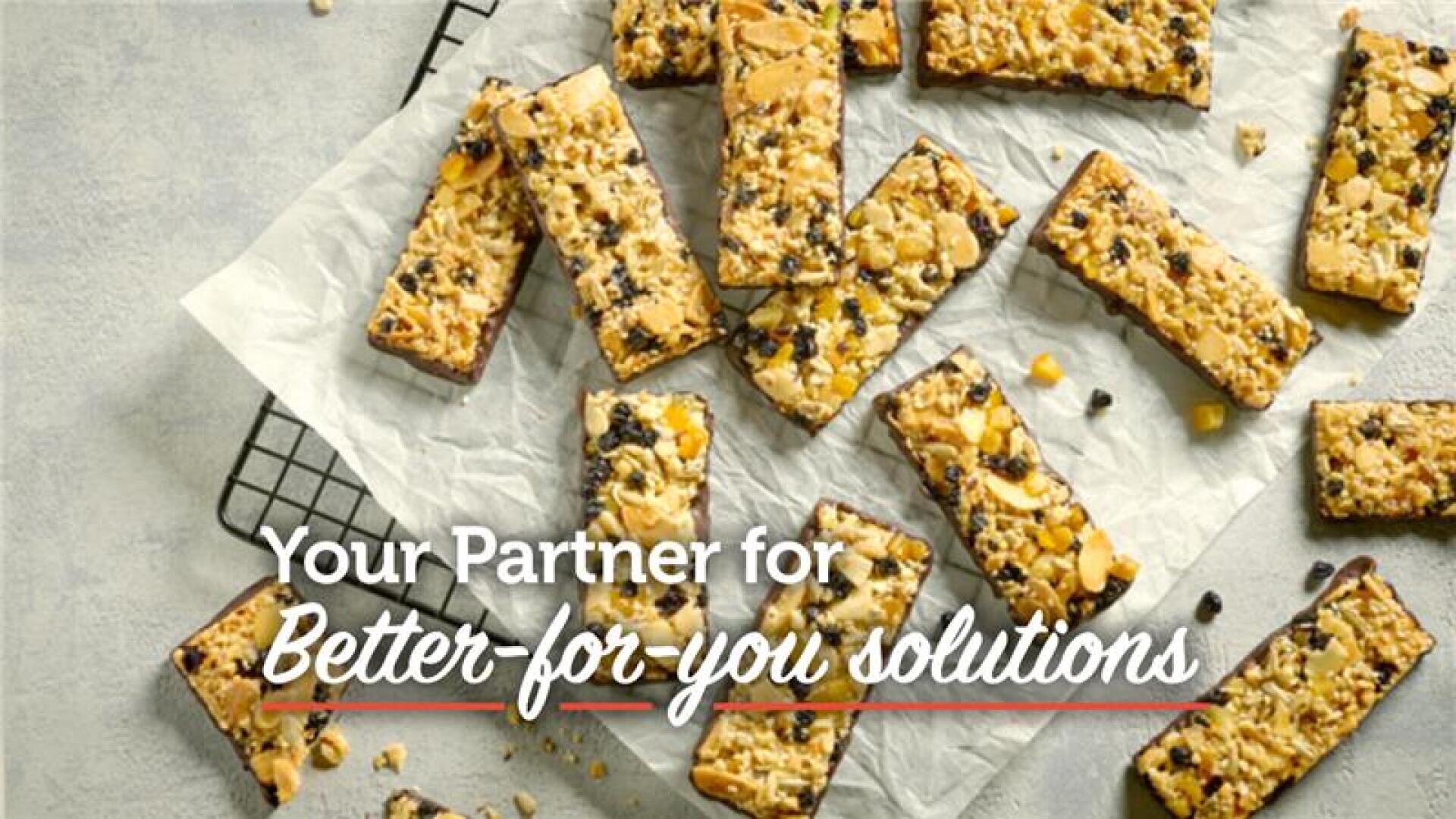 Granola bars with nuts and dried fruit are arranged on parchment paper and a cooling rack. Text on the image reads, Your Partner for Better-for-you solutions.