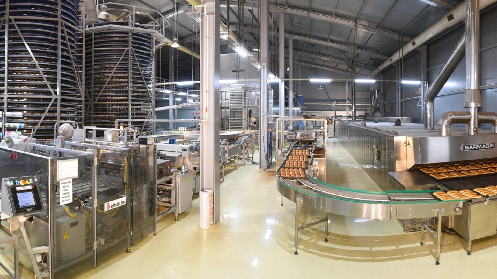 Industrielle Bäckerei mit Förderbändern, die Backwaren transportieren, großen spiralförmigen Kühlregalen, Maschinen und Geräten aus Edelstahl in einer geräumigen, gut beleuchteten Fabrikumgebung.