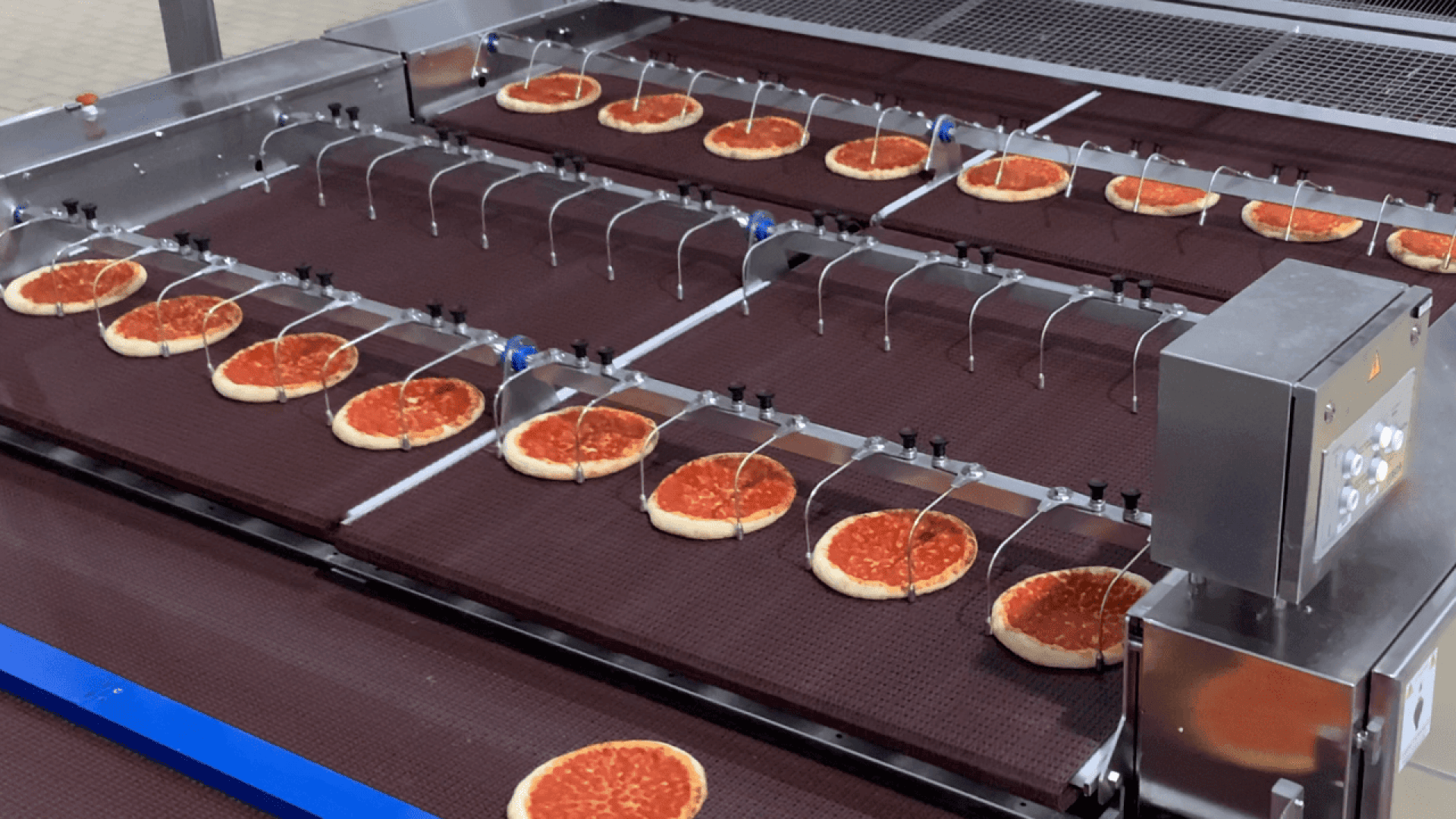 Reihen von Pizzakrusten mit Tomatensoße bewegen sich auf einem Förderband in einer Lebensmittelverarbeitungsanlage unter Maschinen hindurch, die Teil einer automatisierten Pizzaproduktionslinie sind.