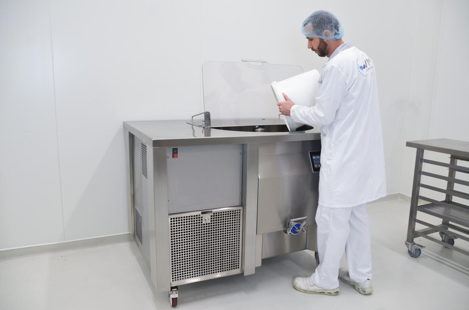 A person in a white lab coat, hair net, and shoe covers pours contents from a container into a large stainless steel industrial machine in a clean, white laboratory or food processing facility.