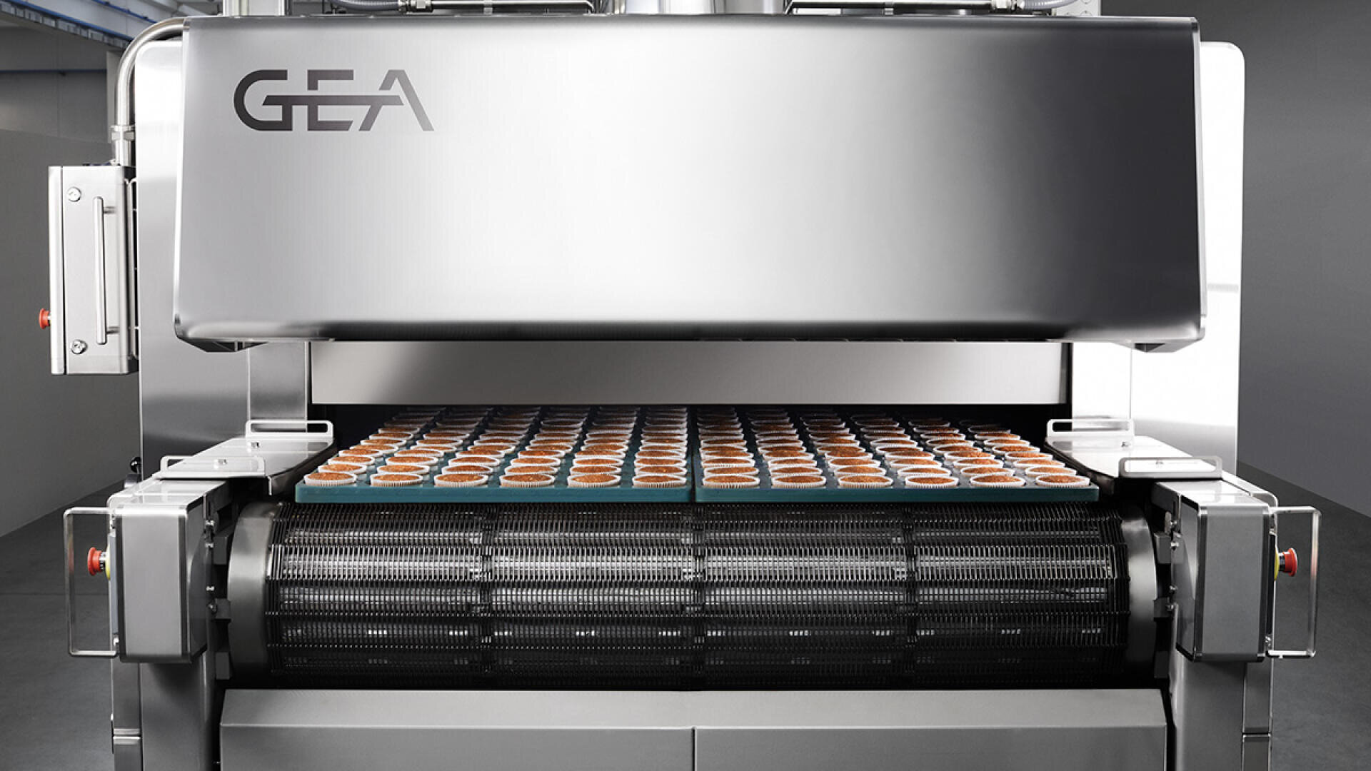 A large industrial food processing machine with the GEA logo dispenses rows of filled baking trays on a conveyor belt in a modern facility.