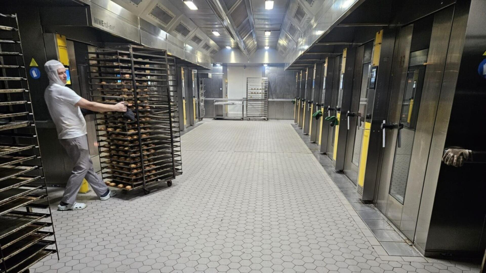 A worker in protective clothing pushes a tall rack of baking trays through a clean, industrial bakery with large ovens lining both sides of the tiled corridor.