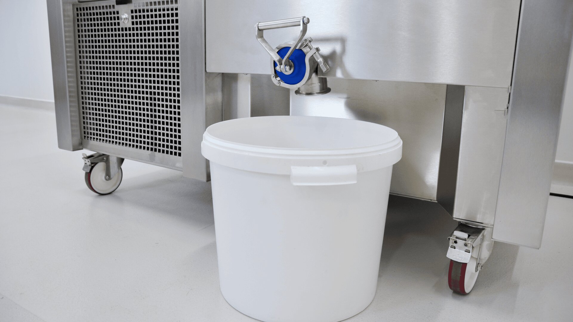 A large white plastic bucket is placed under a metal spout attached to a stainless steel industrial machine on wheels, set on a clean, light-colored floor.