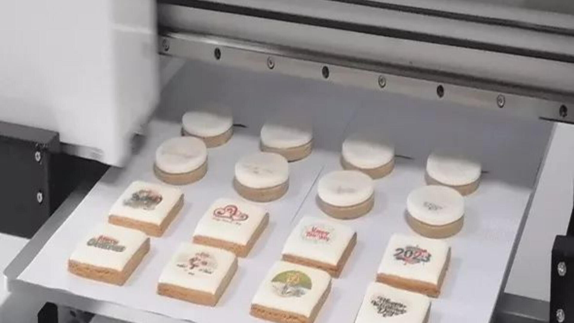 A machine prints colorful designs on square and round cookies arranged in rows on a tray lined with white paper.