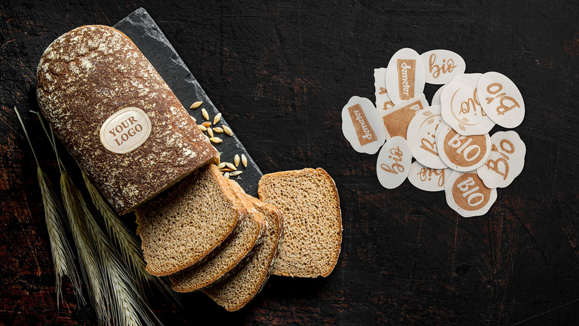 Ein Laib geschnittenes Vollkornbrot mit einem individuellen Logoaufkleber liegt auf einer dunklen Oberfläche neben Weizen, Körnern und einem Stapel runder Aufkleber mit der Aufschrift „Bio“ und „Demeter“ in verschiedenen Schriftarten.
