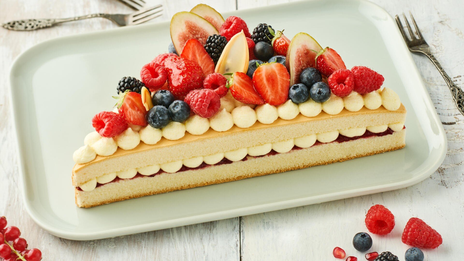 A triangular layered sponge cake topped with piped cream and assorted fresh berries including strawberries, raspberries, blueberries, blackberries, and figs, served on a light-colored rectangular platter.