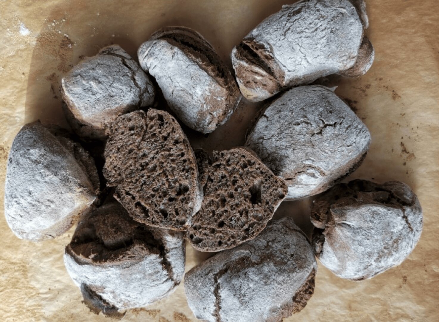 A top-down view of several round, dark brown bread rolls dusted with flour, some of which are cut open to reveal their airy, rustic crumb texture. The rolls are arranged on brown parchment paper.