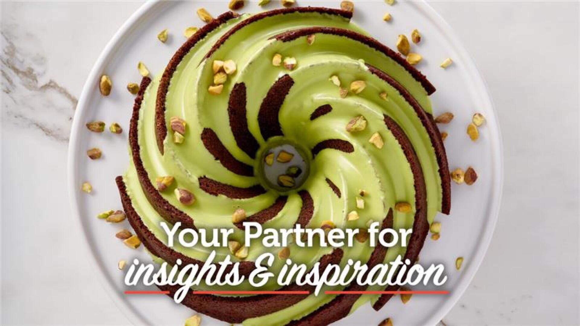 A chocolate bundt cake with green icing and sprinkled pistachios sits on a white plate. Overlaid text reads: Your Partner for insights & inspiration.