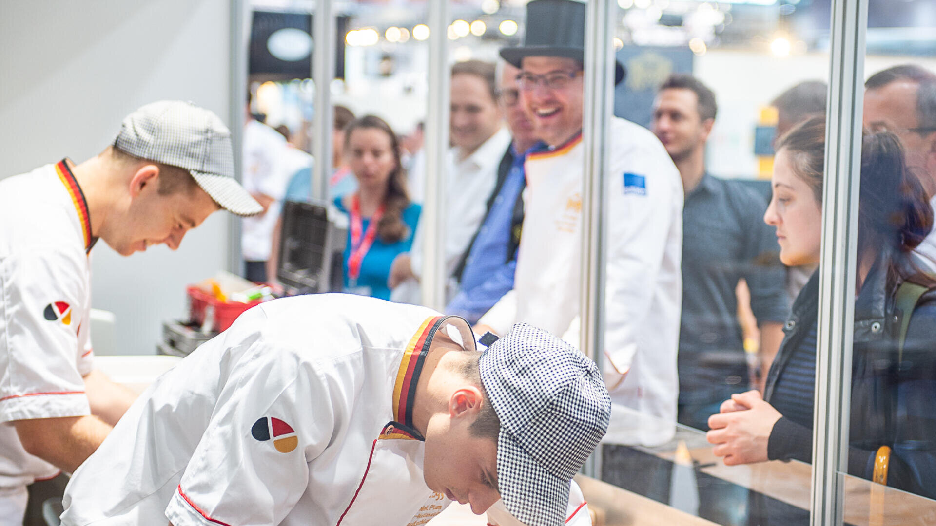 Zwei Köche in weißen Uniformen und Mützen arbeiten hinter einer Glaswand, während eine Gruppe von Personen, darunter ein Mann mit Zylinder und Laborkittel, sie bei einer Veranstaltung oder einem Wettbewerb beobachtet.