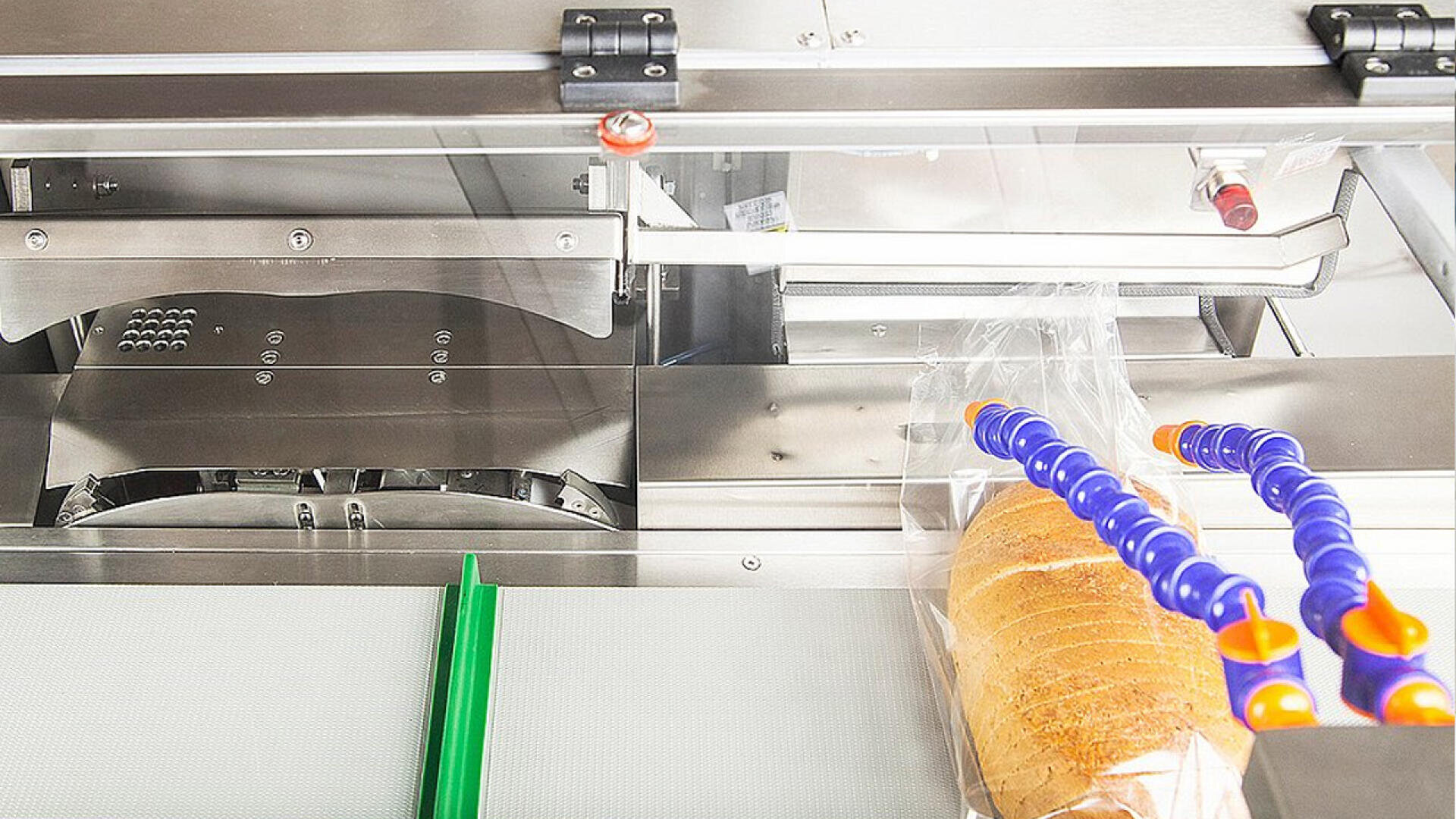 A loaf of sliced bread in a clear plastic bag is being processed by an automated packaging machine with metal and plastic components, including blue and orange flexible tubes.