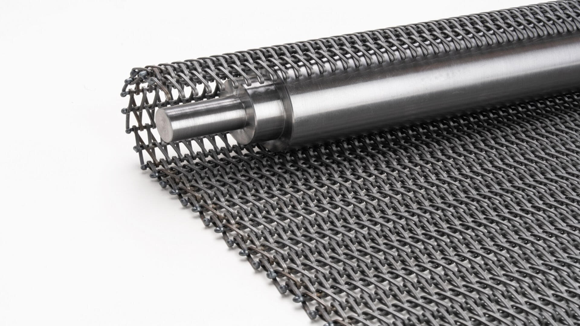 A close-up of a metal conveyor belt with a cylindrical metal roller, partially unrolled, showing an interwoven mesh pattern on a white background.