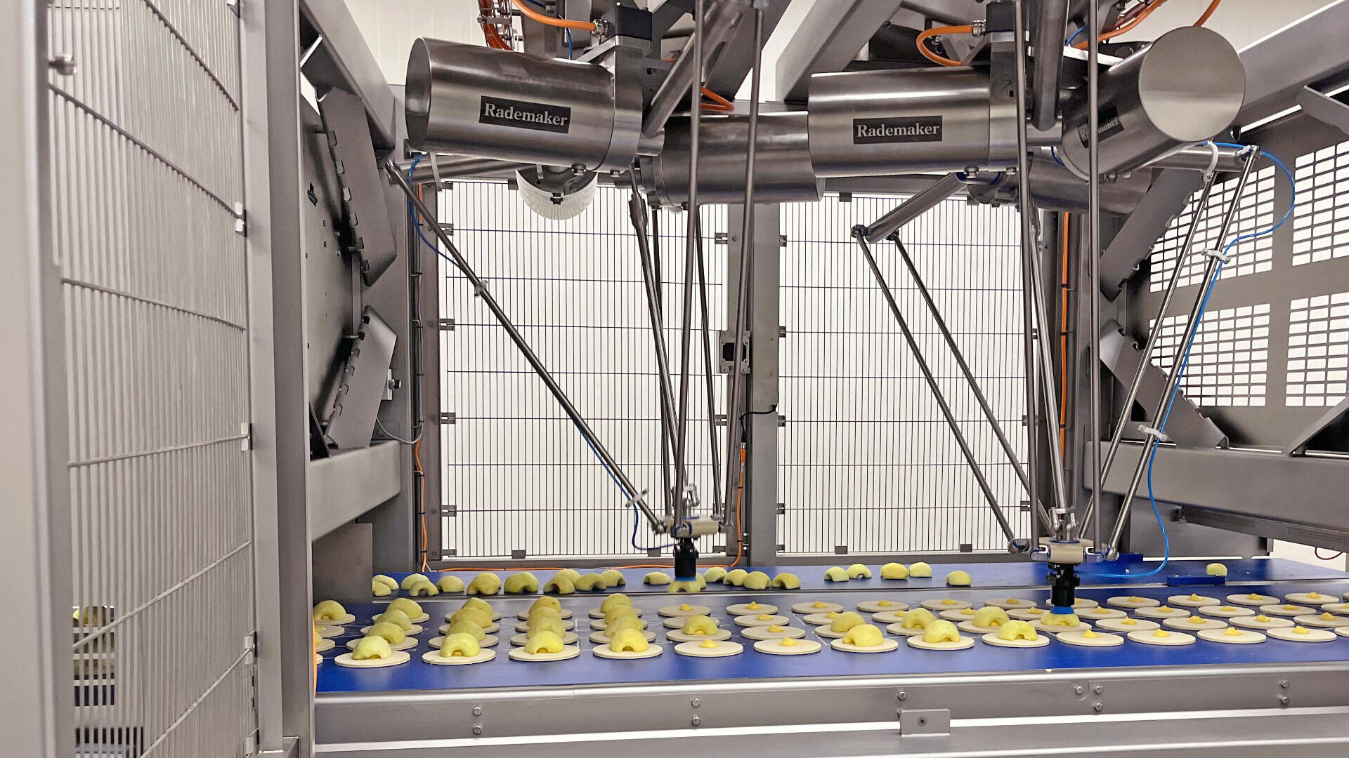 A food processing machine with robotic arms placing dough pieces onto a conveyor belt inside a factory. The background features metal fencing and equipment.