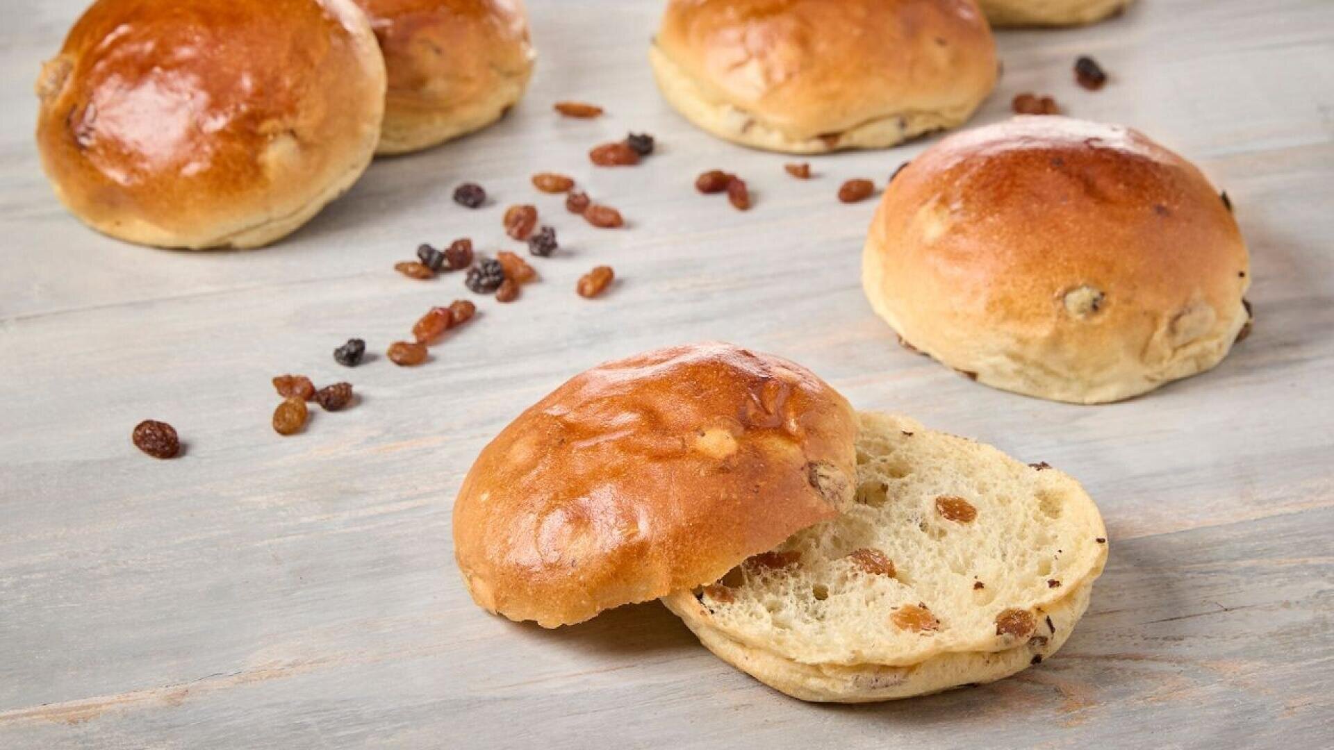 Several shiny, golden-brown raisin buns are spread on a light wooden surface, with one bun cut open to reveal its soft, fluffy interior filled with raisins. Loose raisins are scattered around the buns.