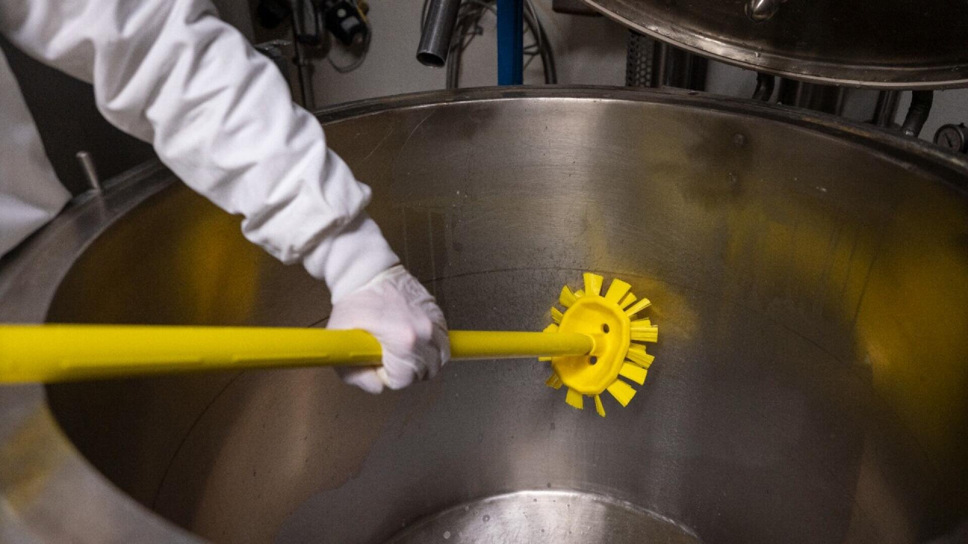 A person wearing a white glove uses a yellow brush with a long handle to clean the inside of a large stainless steel container.