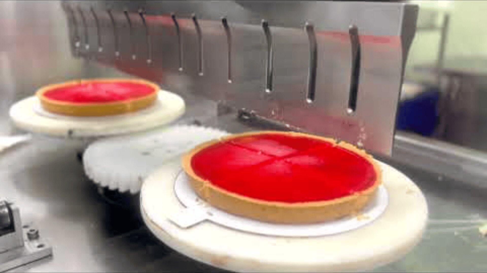 Zwei rote Torten auf runden Tabletts befinden sich auf einem Förderband, wobei eine Torte von einer Metallschneidemaschine in vier Stücke geschnitten wird.