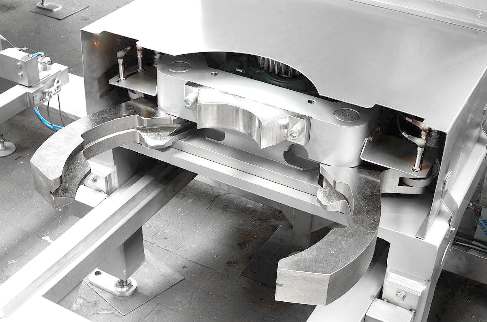 Close-up of a large, metallic industrial machine part with curved, claw-like arms and various bolts and components, situated on a factory floor.