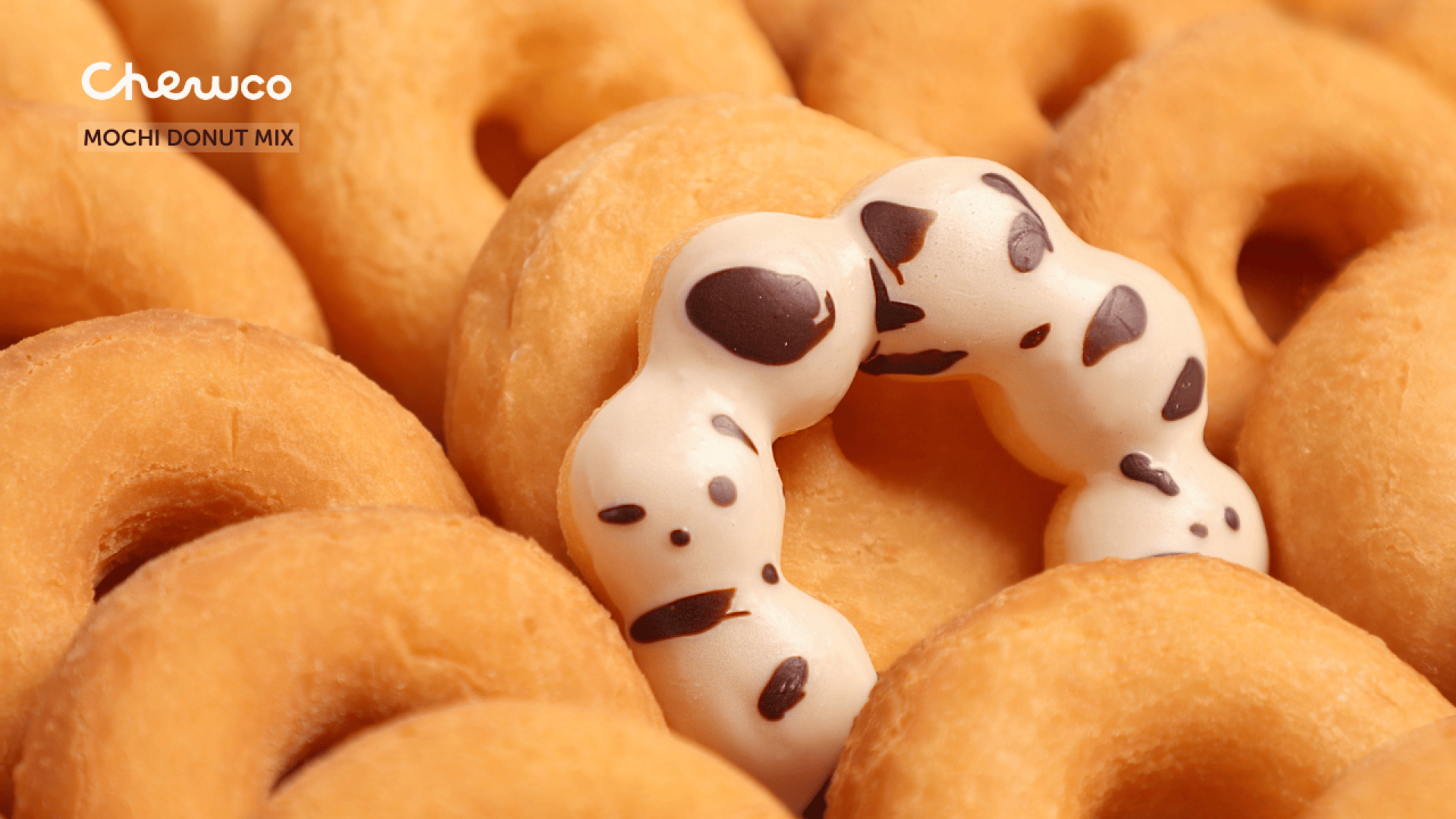 A group of plain glazed donuts surrounds a unique white mochi donut with chocolate spots, forming a contrast in color and shape. The text Chewco MOCHI DONUT MIX appears in the top left corner.
