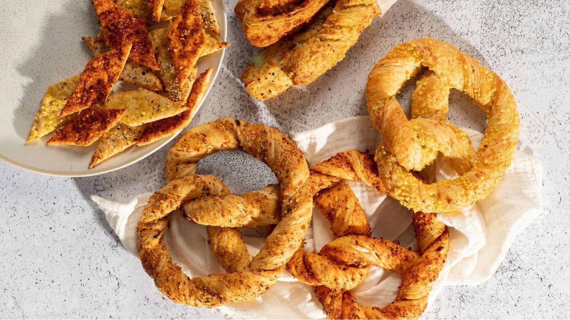 Three large, golden-brown pretzels and twisted breadsticks are arranged on a white cloth, with a plate of thin, crispy bread pieces on the side, all set on a light-colored textured surface.