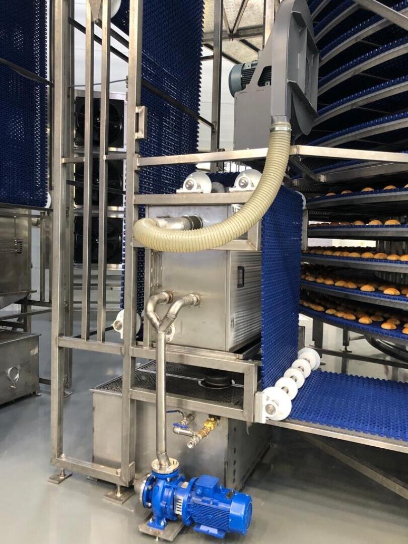 A stainless steel industrial food processing machine with blue conveyor belts carrying rows of round baked goods, metal piping, a blue motor pump, and a vent hose in a clean factory environment.