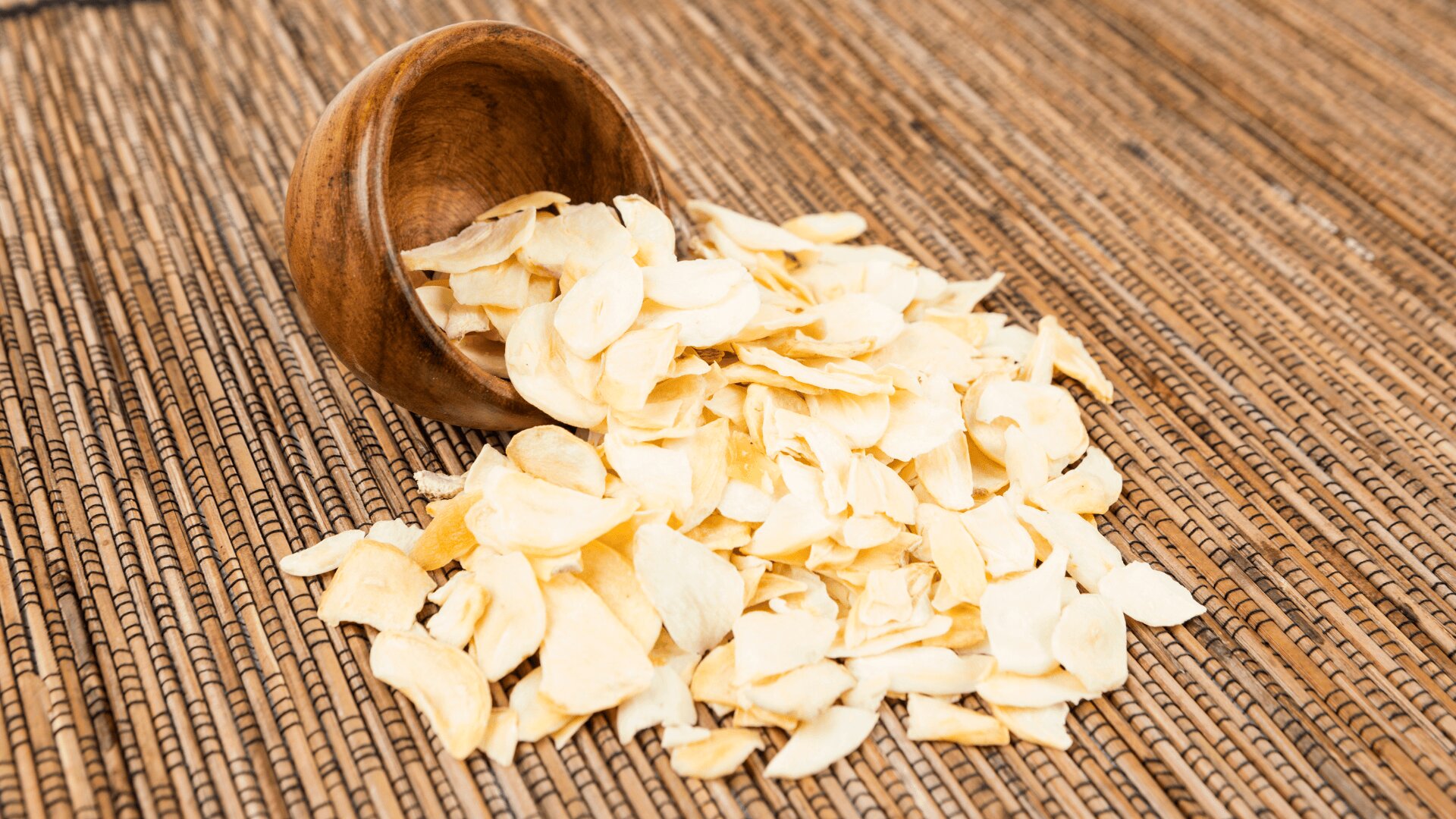 Thinly sliced dried garlic pieces spill out of a small wooden bowl onto a woven straw mat.