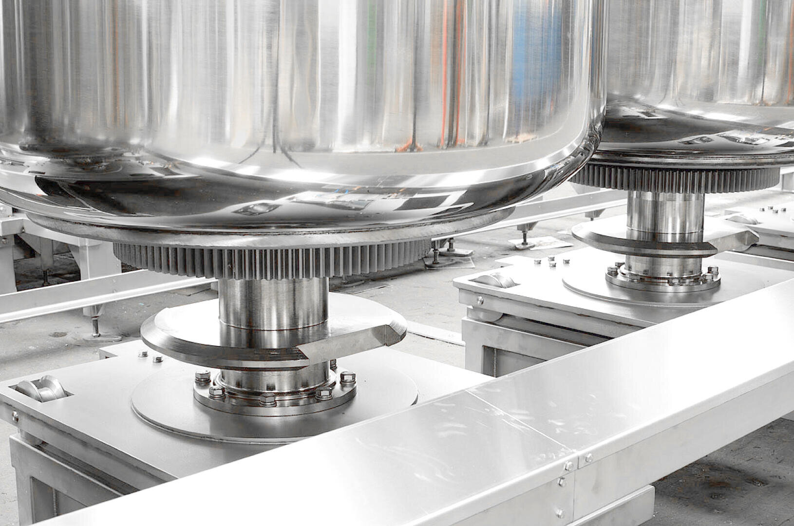 Close-up of large, shiny stainless steel industrial tanks with visible metal bases and gears, situated indoors on a clean, gray factory floor. The image emphasizes modern equipment and clean manufacturing.