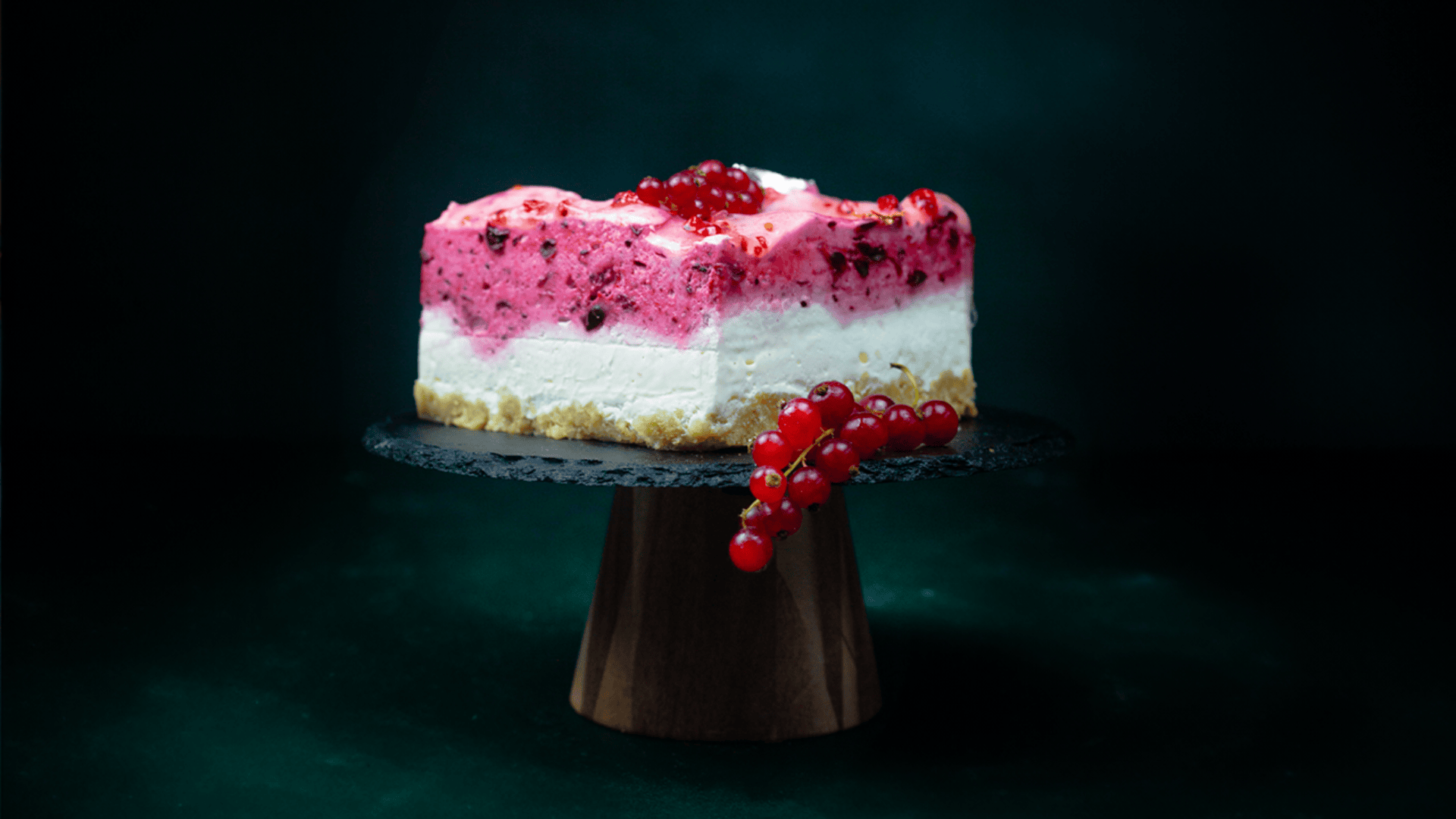 A slice of layered berry and cream cheesecake sits on a black cake stand, garnished with red currants, against a dark green background.