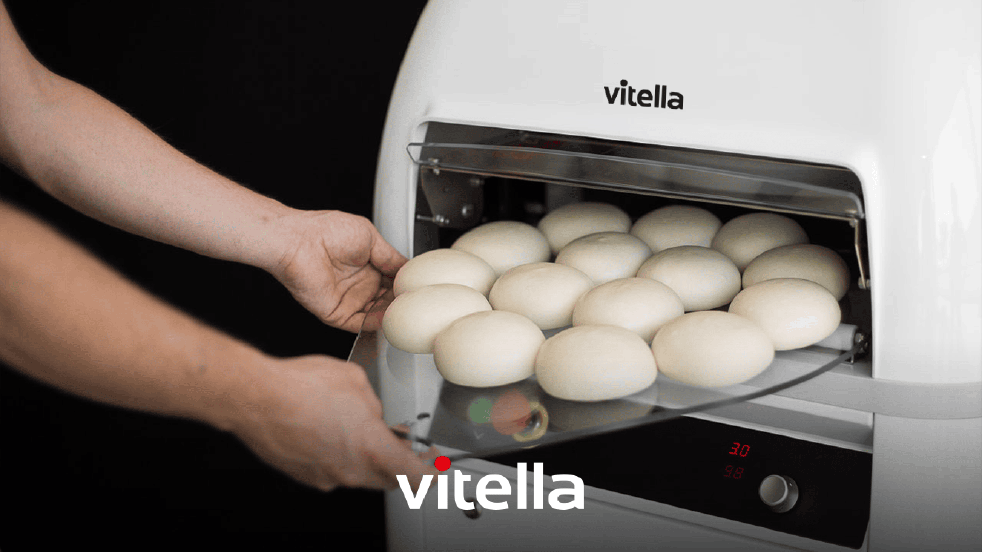 A person places a tray of round dough balls into a white Vitella dough processing machine, with the Vitella logo visible on the front.