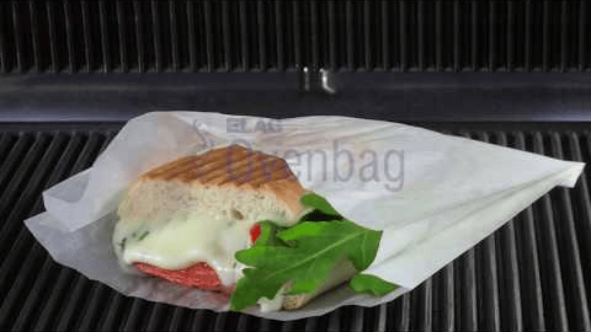 A sandwich with melted cheese, slices of meat, and fresh arugula is wrapped in white paper and placed on a grill or panini press. The sandwich appears partially toasted.