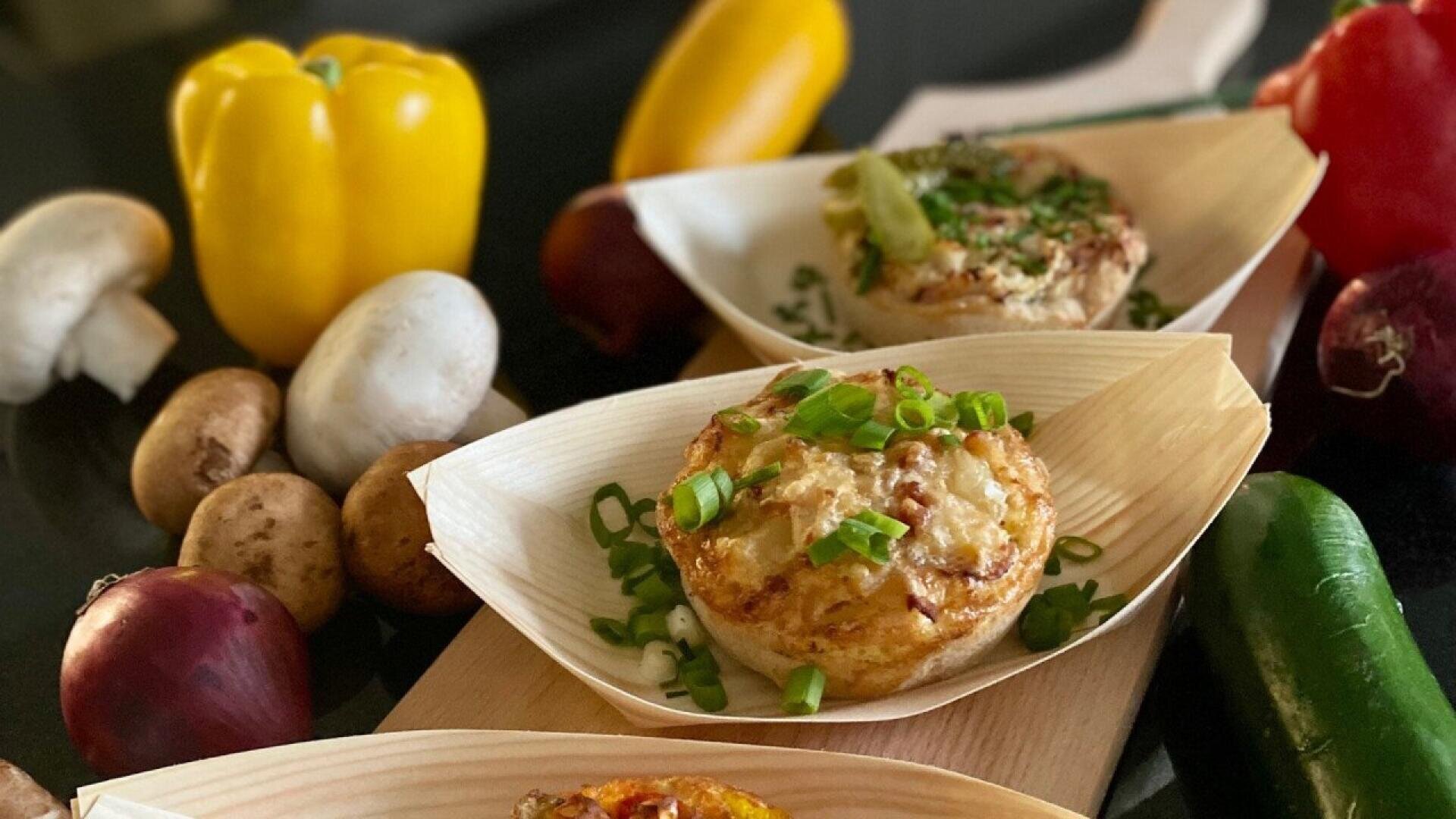 Three individual egg muffins garnished with chopped green onions are served in small wooden trays. Surrounding them are fresh vegetables including mushrooms, yellow and red bell peppers, onions, and a jalapeño.