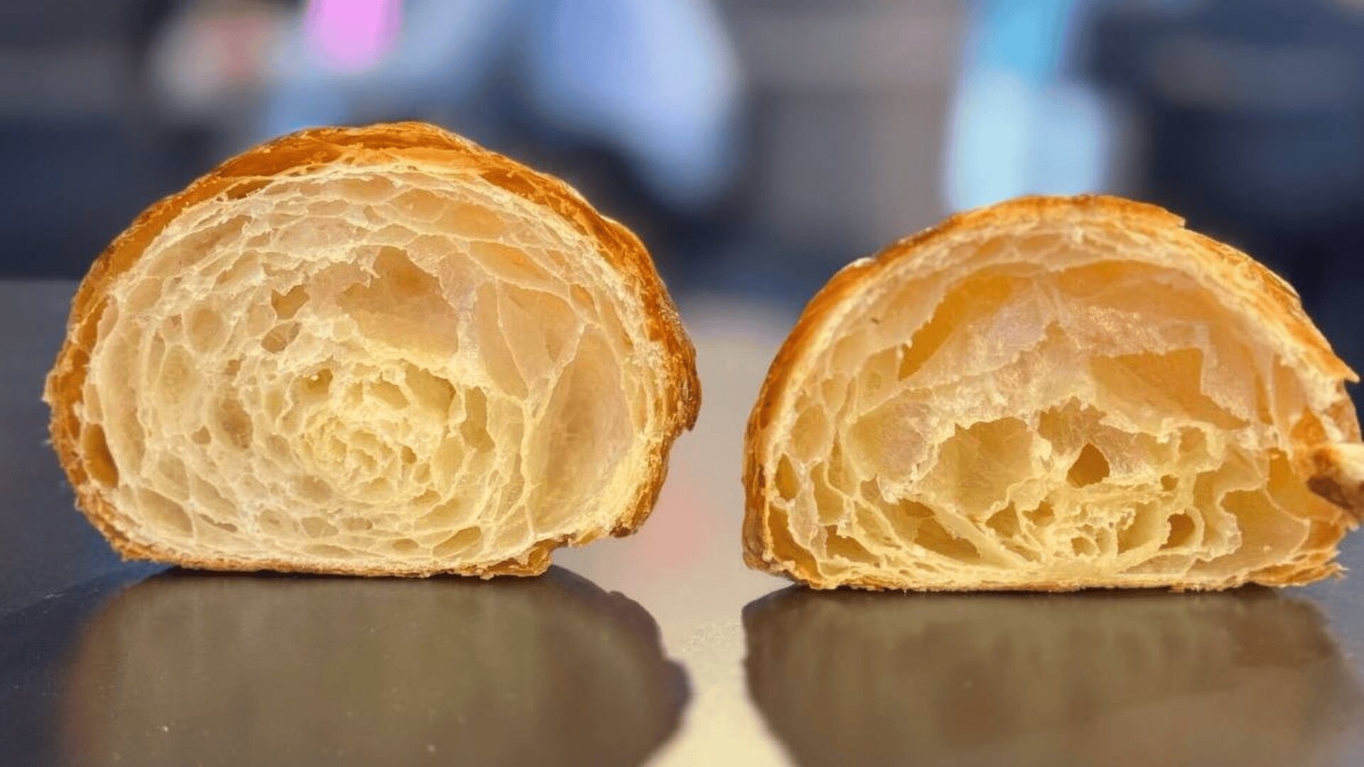 Two halves of a croissant are placed upright on a surface, showing their airy, flaky interior layers and golden-brown crust. The background is blurred.