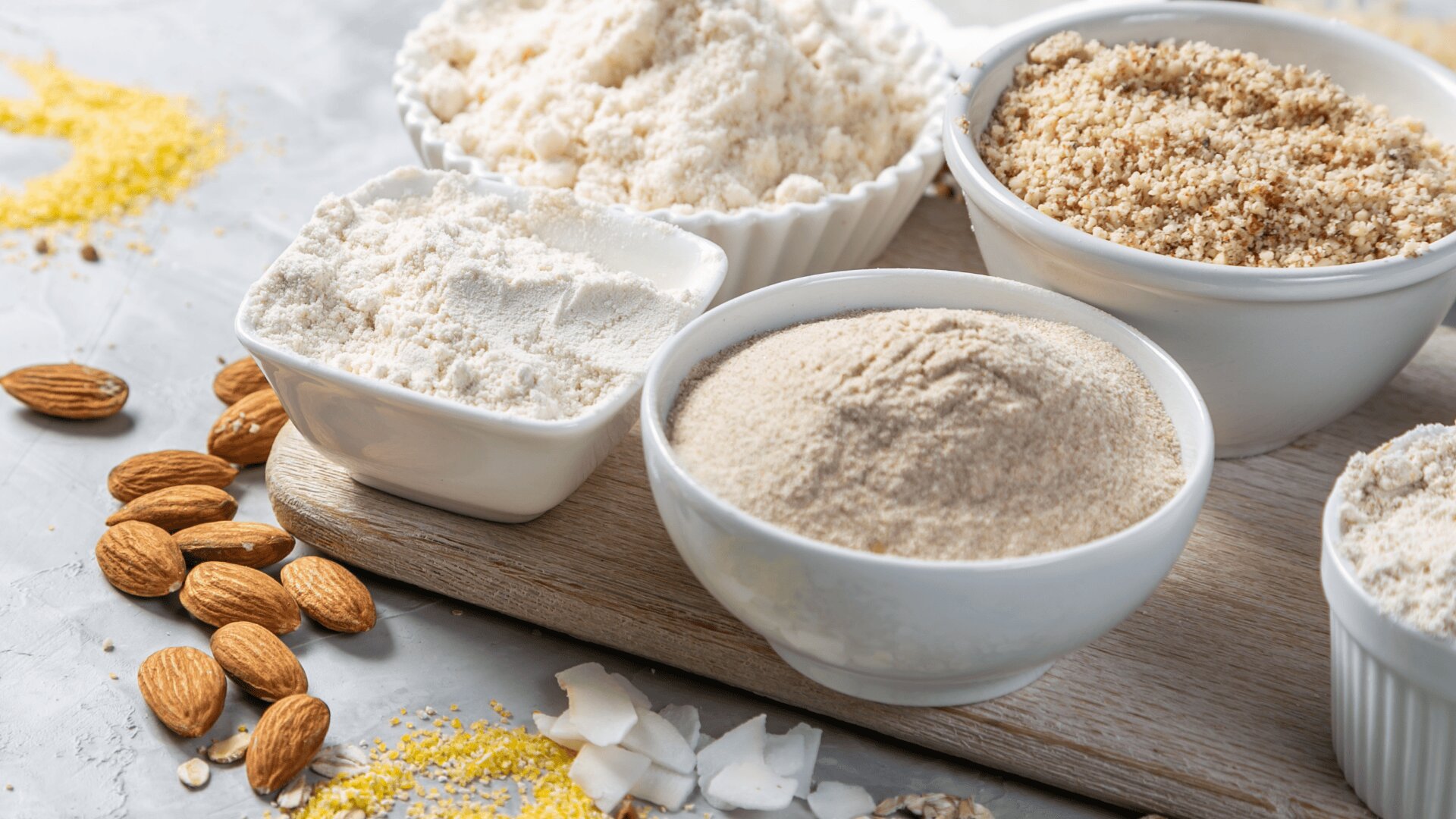 Auf einem Holzbrett sind Schälchen mit verschiedenen glutenfreien Mehlen und gemahlenen Nüssen angeordnet, umgeben von ganzen Mandeln, Kokosraspeln und Mehl, das auf eine hellgraue Fläche gestreut wird.