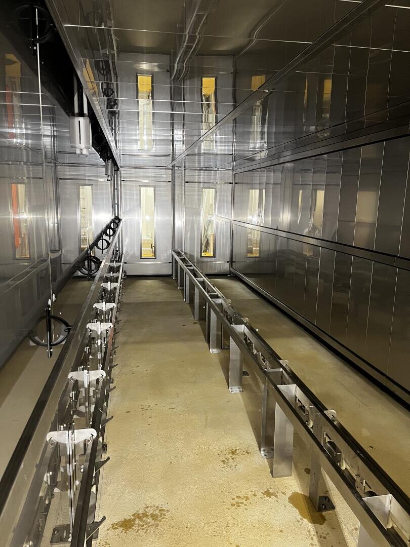 Ein hell erleuchteter, industrieller Metallraum mit reflektierenden Wänden, einem beigen Boden und einem langen Metallrahmen, der parallel zum Boden verläuft, möglicherweise für die Installation oder Montage von Geräten.
