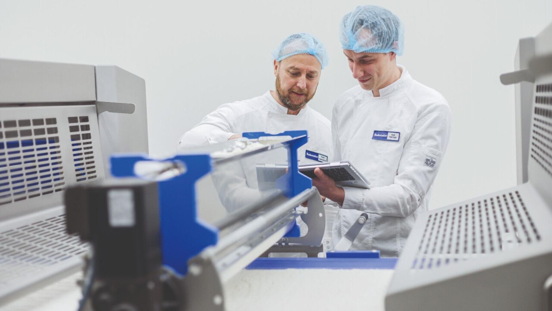 Zwei Arbeiter in weißen Uniformen und blauen Haarnetzen bedienen Maschinen in einer sauberen, industriellen Umgebung. Einer zeigt auf die Geräte, der andere hält ein Tablet in der Hand, beide sind auf ihre Aufgaben konzentriert.