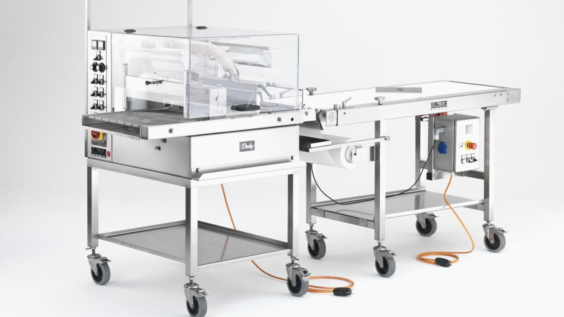 A stainless steel commercial packaging machine with wheels, control panels, a conveyor belt, and protective clear shields, set against a plain white background with visible orange power cords.