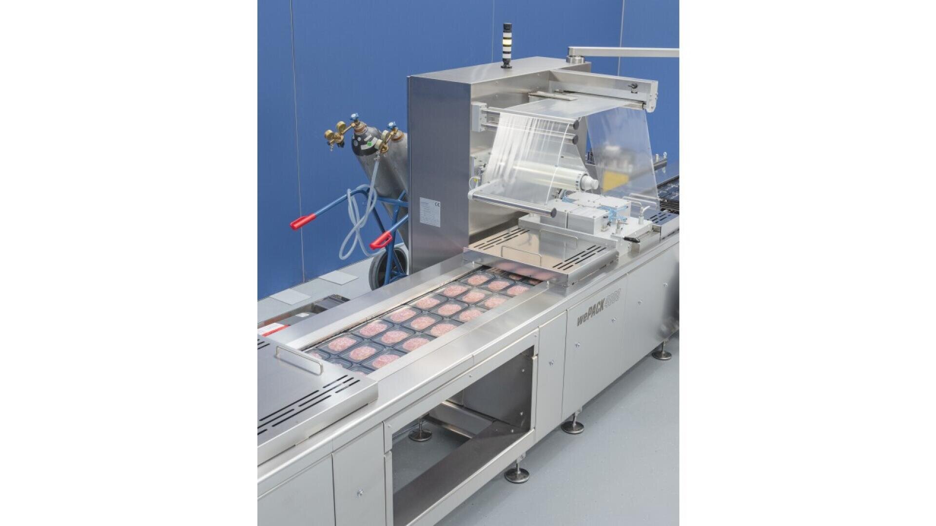 A food packaging machine with sealed trays containing food products, likely meat patties, moving along a conveyor belt in a clean, industrial setting with blue walls and various equipment nearby.