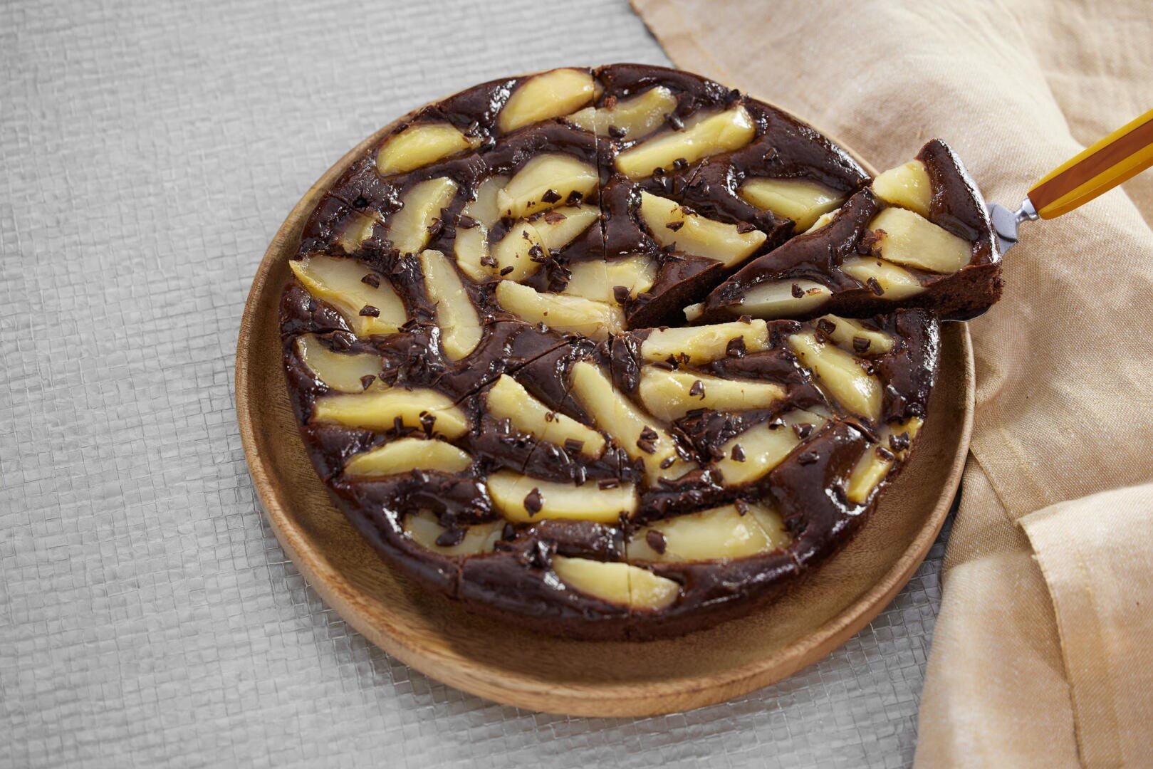 Ein runder Schokoladenkuchen, belegt mit Birnenscheiben, auf einer Holzplatte ausgestellt. Eine Scheibe wird mit einem Tortenheber angehoben. Neben dem Teller liegt ein beiges Tuch auf einer hellen, strukturierten Oberfläche.