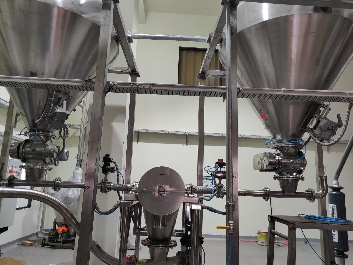 Two large stainless steel industrial hoppers with conical bottoms are connected by pipes and valves in a clean, indoor processing facility. Various wires, tubes, and machinery surround the equipment.