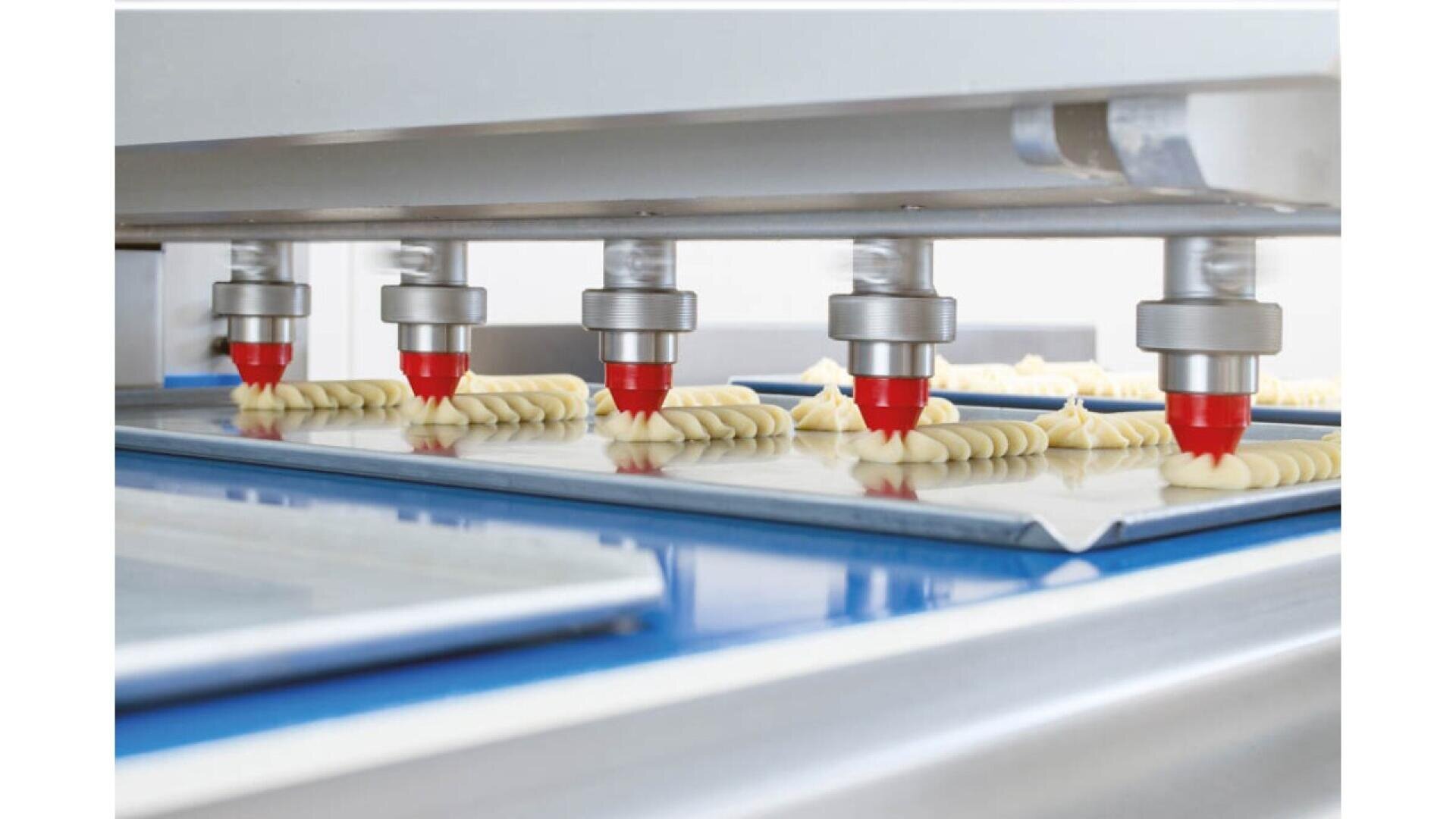 Fünf automatisierte Düsen spritzen Teig auf Backbleche in einer Lebensmittelproduktionslinie und erzeugen so in einer industriellen Bäckerei gleichmäßig geformte Portionen.