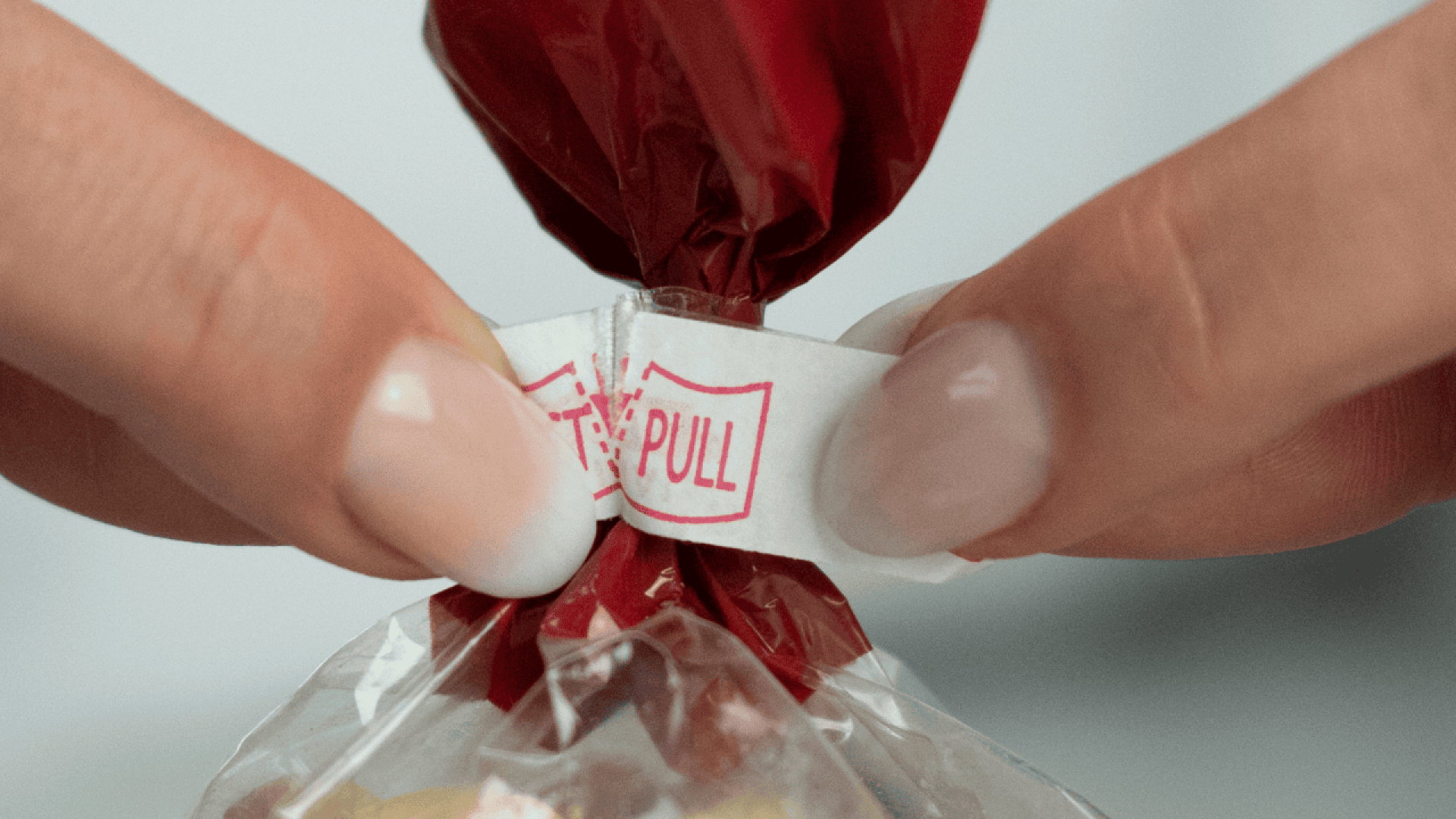 Nahaufnahme von Händen, die eine Plastiktüte öffnen, indem sie an einer weiß-roten "PULL"-Lasche um das gedrehte Ende der Tüte ziehen.