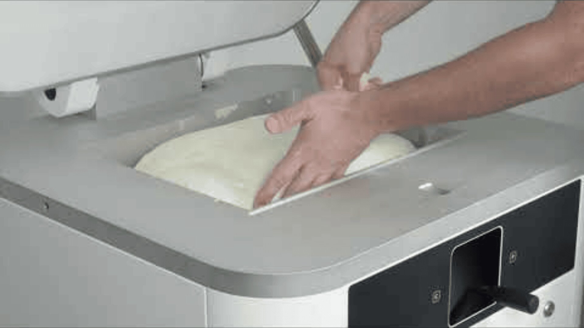 Eine Person knetet Teig in einer Teigknetmaschine und drückt den Teig mit beiden Händen. Der Deckel der Maschine ist geöffnet und zeigt den Teig und die Hände des Arbeiters.
