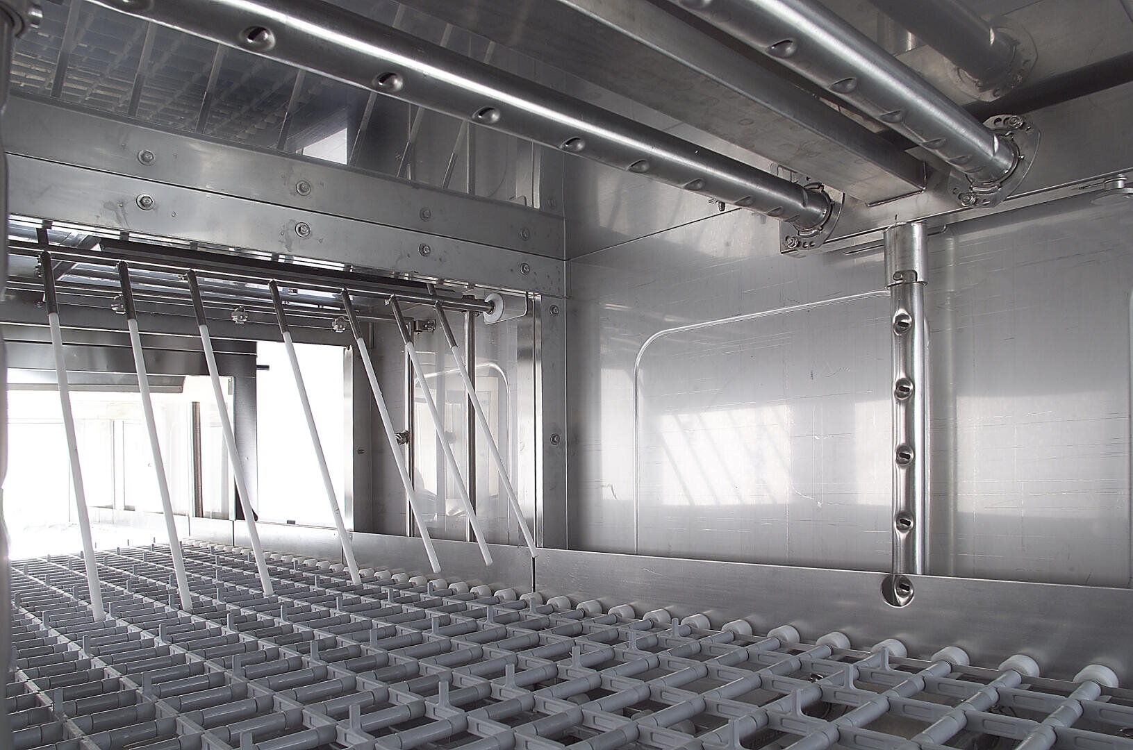 Close-up view of the inside of an industrial dishwasher, showing metal racks, spray arms, and nozzles, all made of stainless steel, designed for cleaning trays and kitchen equipment.