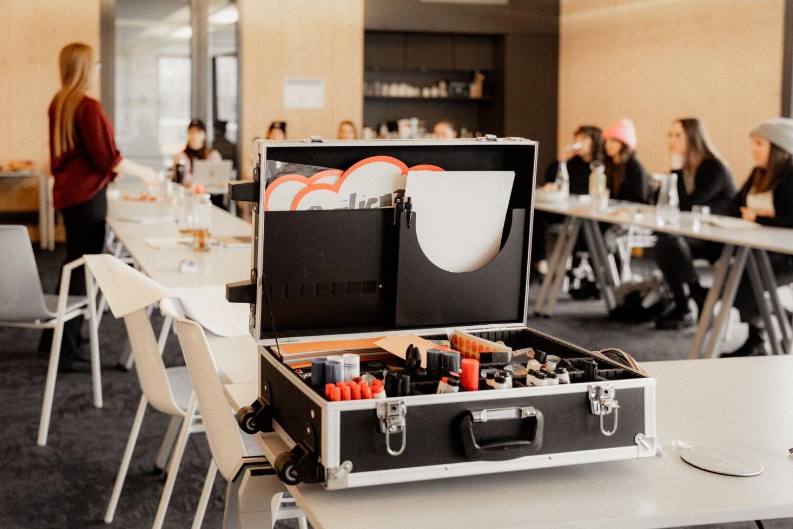 Ein Koffer, gefüllt mit Stiften und Büromaterial, steht offen auf einem Tisch in einem modernen Konferenzraum, in dem Menschen um Tische herum sitzen und jemand an der Stirnseite des Raumes steht.