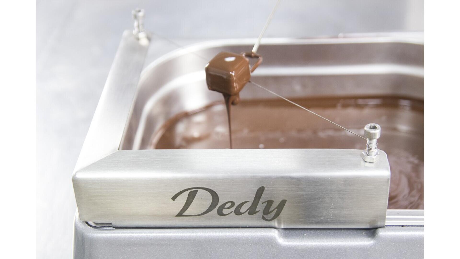 A chocolate-covered confection is suspended on a thin wire above a pan filled with melted chocolate. The metal machine has Dedy engraved on the front.