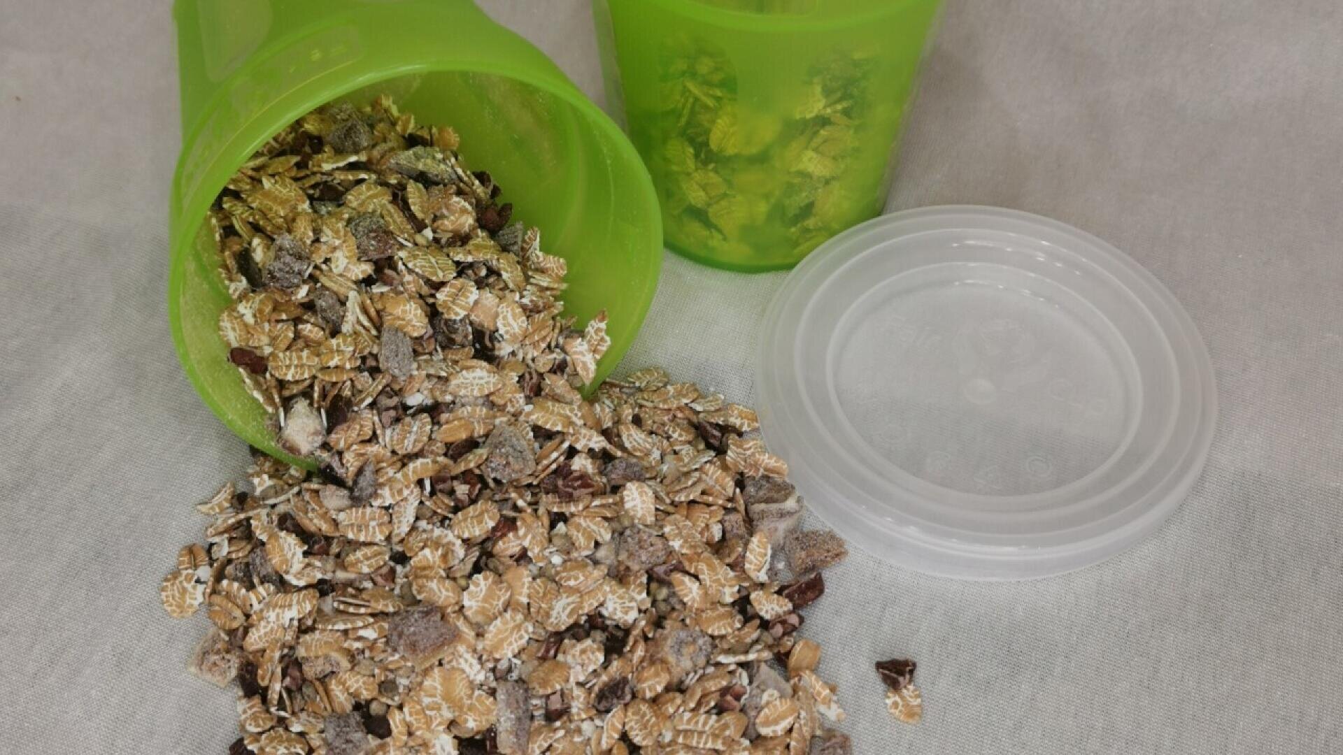 A green plastic container tipped over, spilling a mixture of dry muesli or cereal flakes onto a white surface. Another green container and a clear lid are nearby.