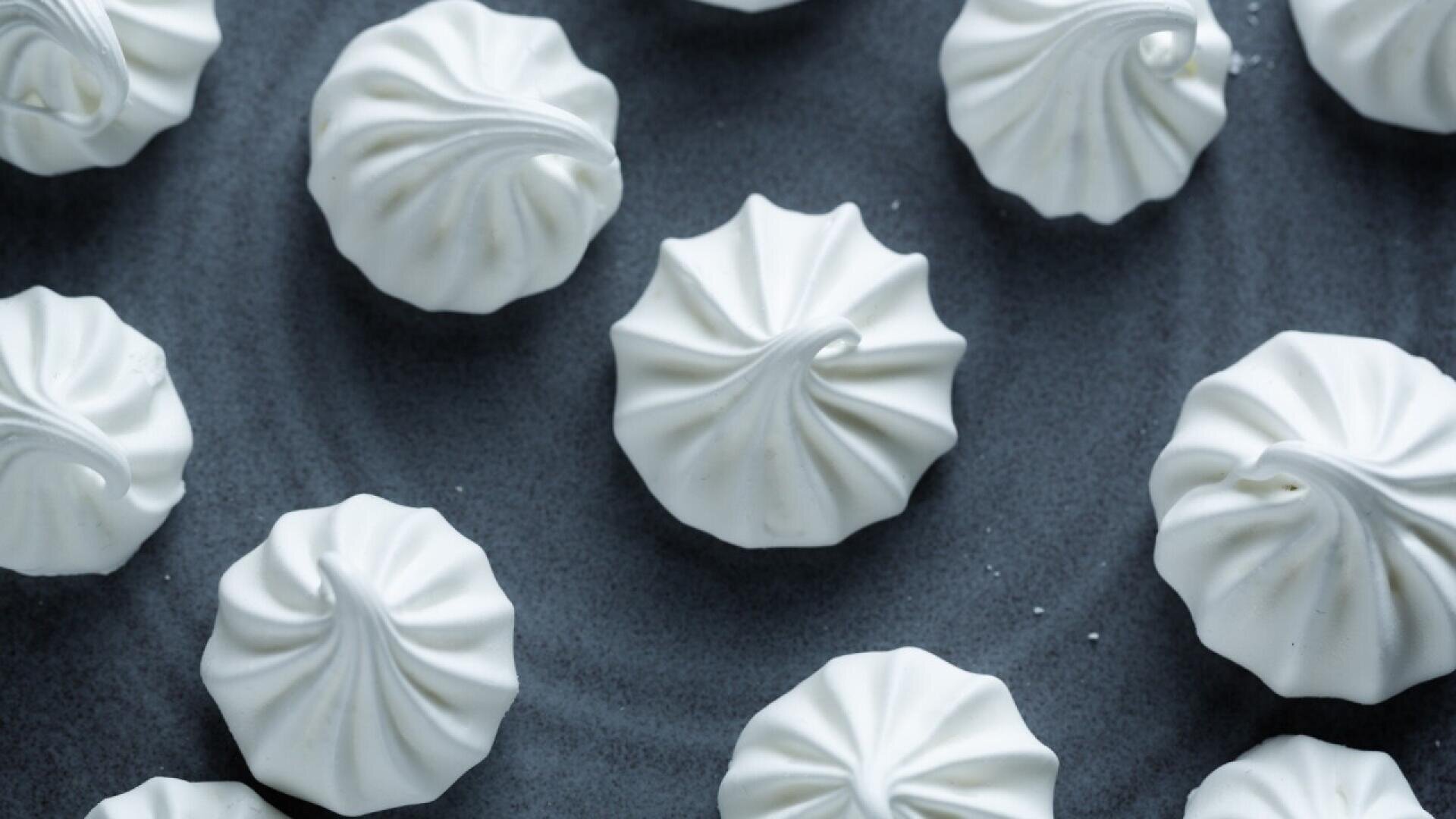 Close-up of white meringue cookies arranged on a dark gray surface. Each meringue has a swirled, ridged pattern and a glossy finish.