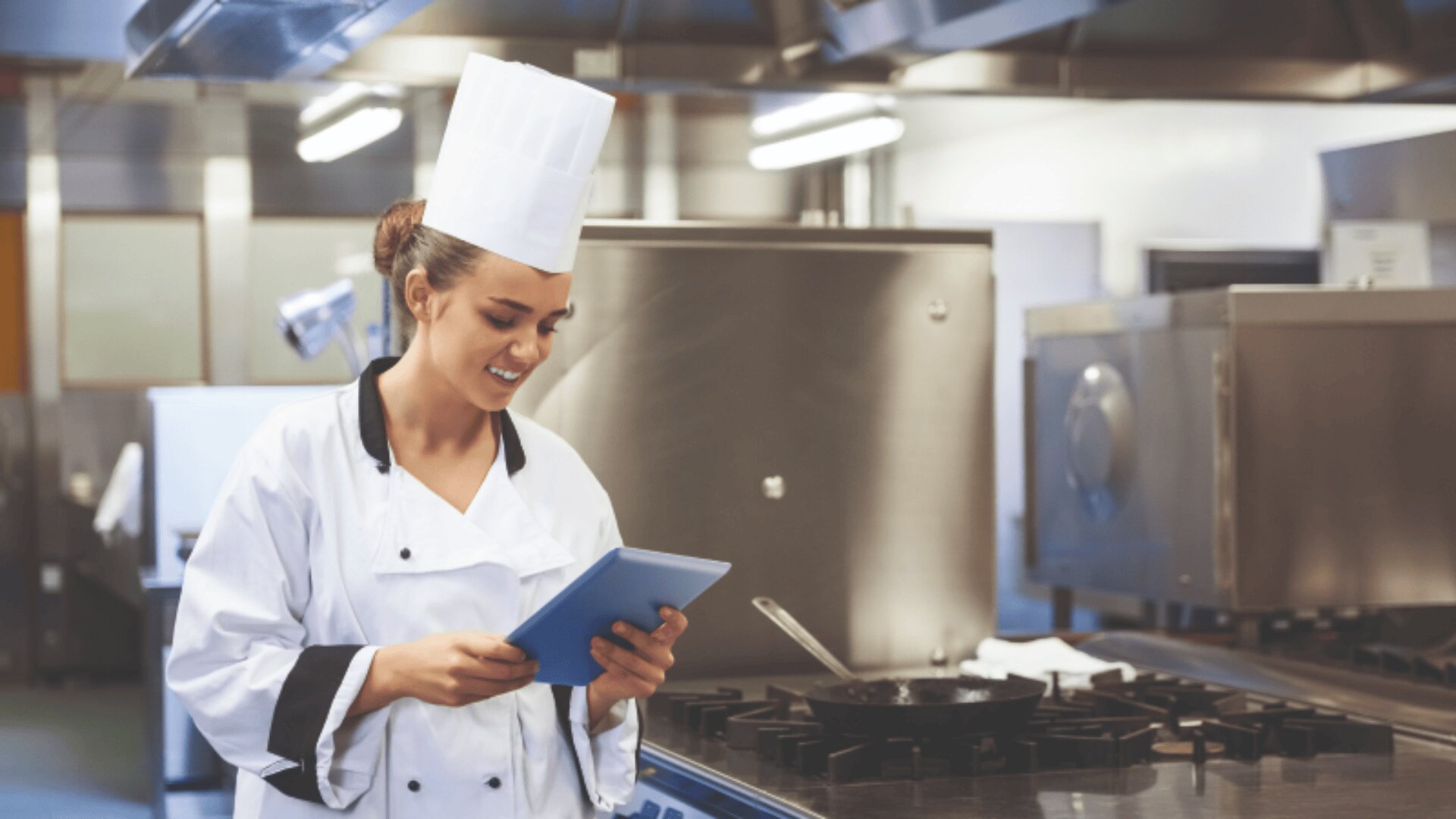 A chef in a white uniform and hat stands in a commercial kitchen, smiling while looking at a tablet. Stainless steel appliances and a stovetop are visible in the background.