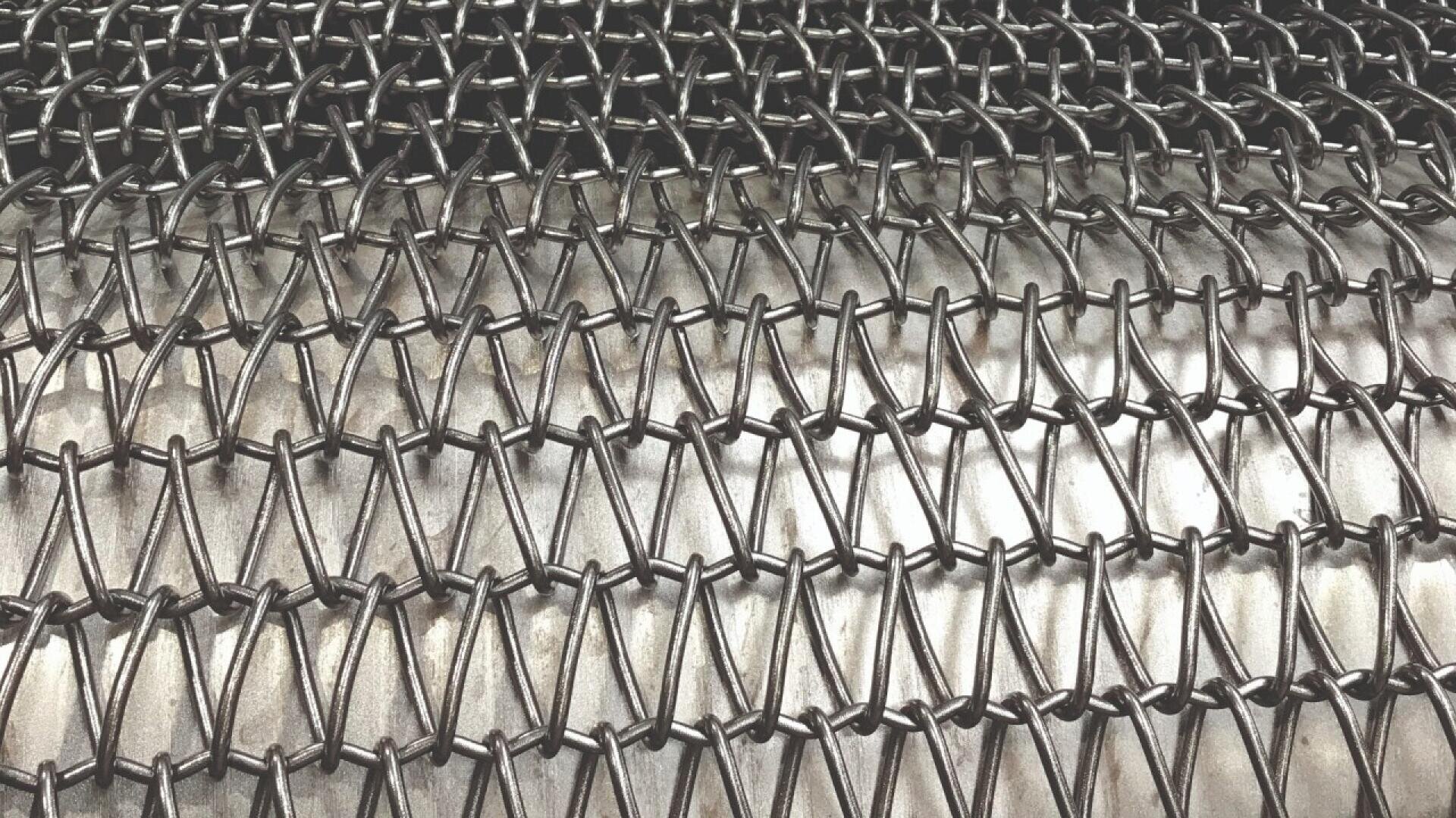 Close-up of a tightly woven metal wire mesh with a zigzag pattern, creating a repeating geometric design. The mesh lies flat on a metallic surface, highlighting the texture and precision of the interlocking wires.