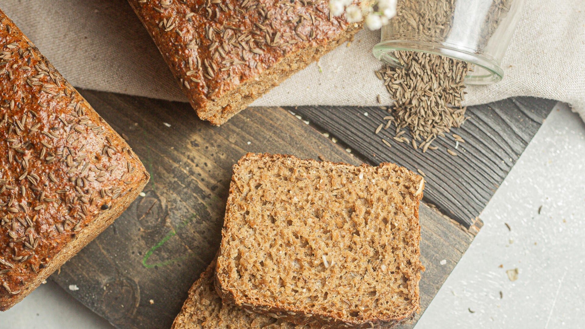 Slices and loaves of whole grain bread with caraway seeds on top, placed on a wooden board. A glass jar with spilled caraway seeds and a beige cloth are also visible.
