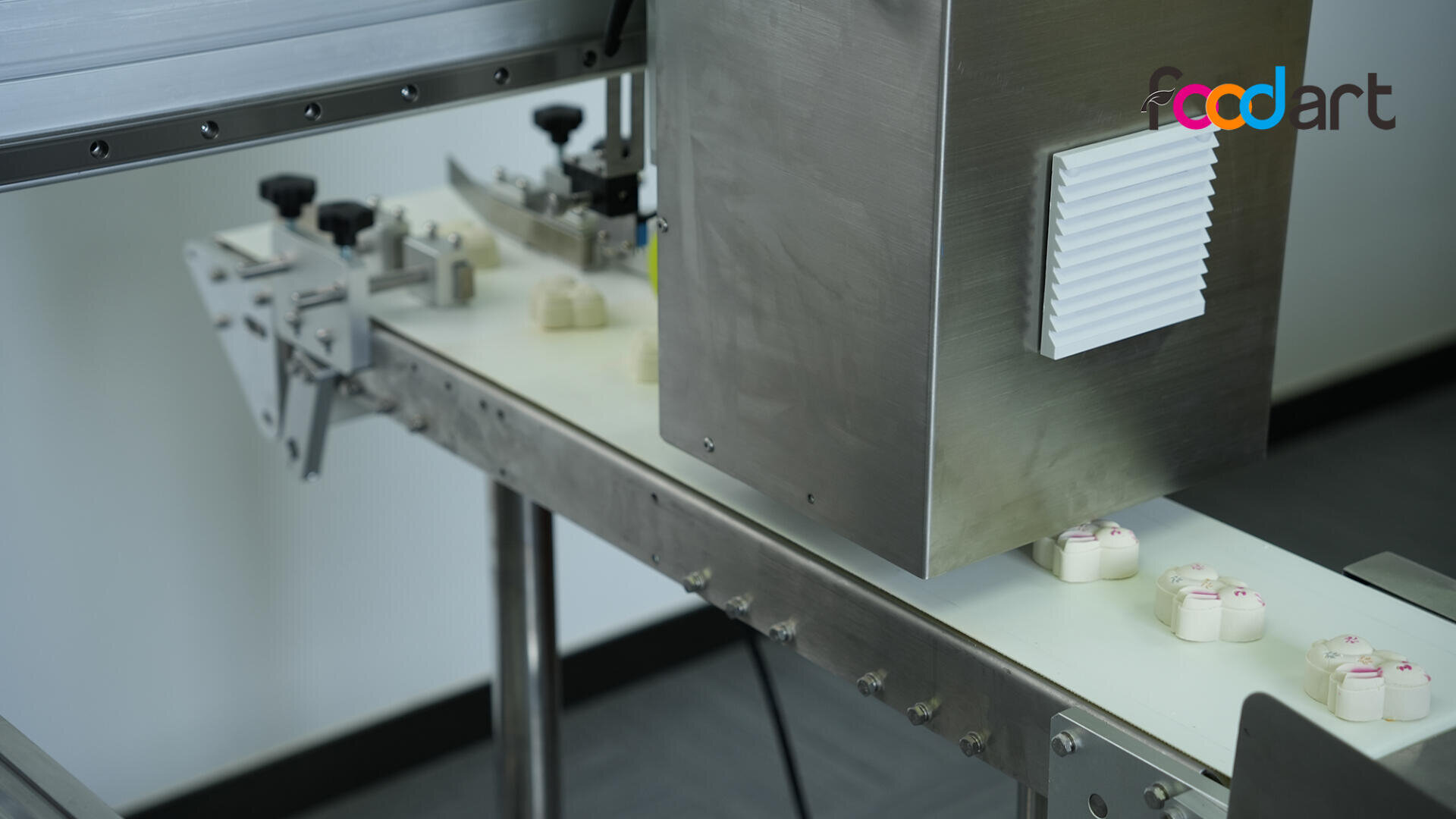 Small white food items with pink decorations move along a conveyor belt toward a stainless steel inspection or packaging machine, under bright lighting. The foodart logo is visible on the top right corner.